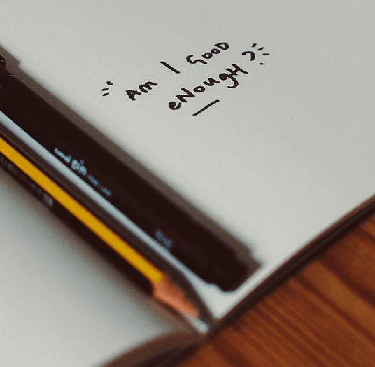 Journal open with a pen and marker resting on a page that reads "am I good enough" in hand written lettering by someone dealing with imposter syndrome